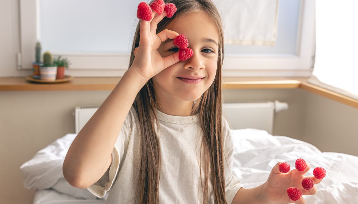 Funny little girl with raspberries on her fingers in bed in the morning
