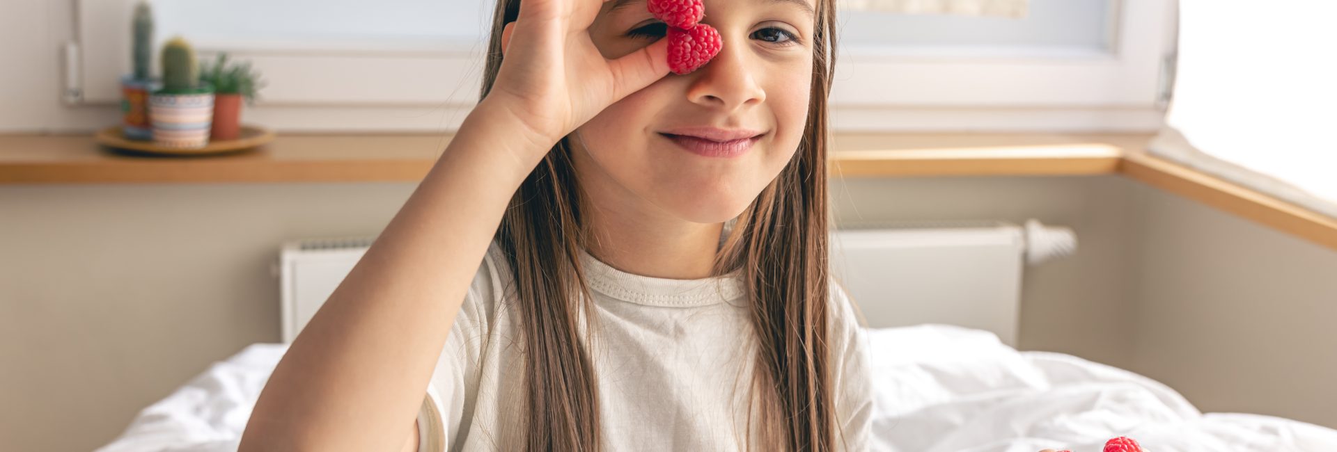 Funny little girl with raspberries on her fingers in bed in the morning